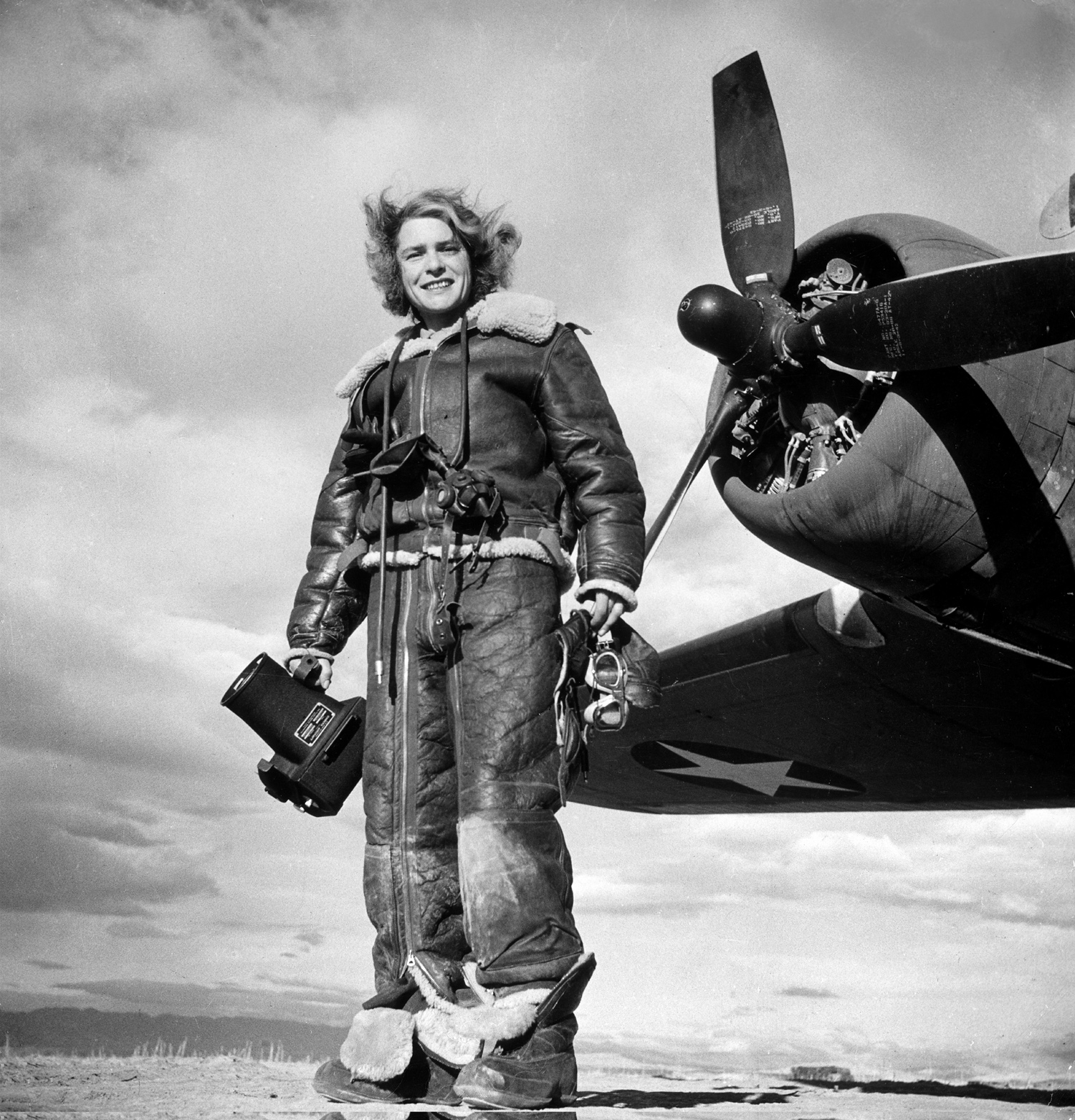 LIFE photographer Margaret Bourke-White clad in fleece flight suit while holding aerial camera, standing in front of Flying Fortress bomber in which she made combat mission photographs of the US attack on Tunis.